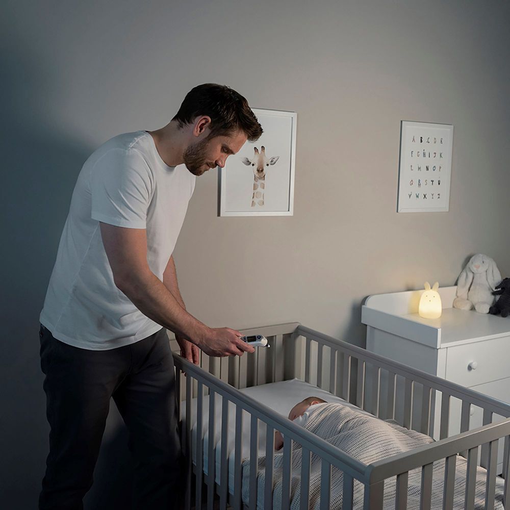 Mann misst Temperatur eines Babys im Bett mit Braun ThermoScan 7+. Umgebung: Kinderzimmer mit Babybett.