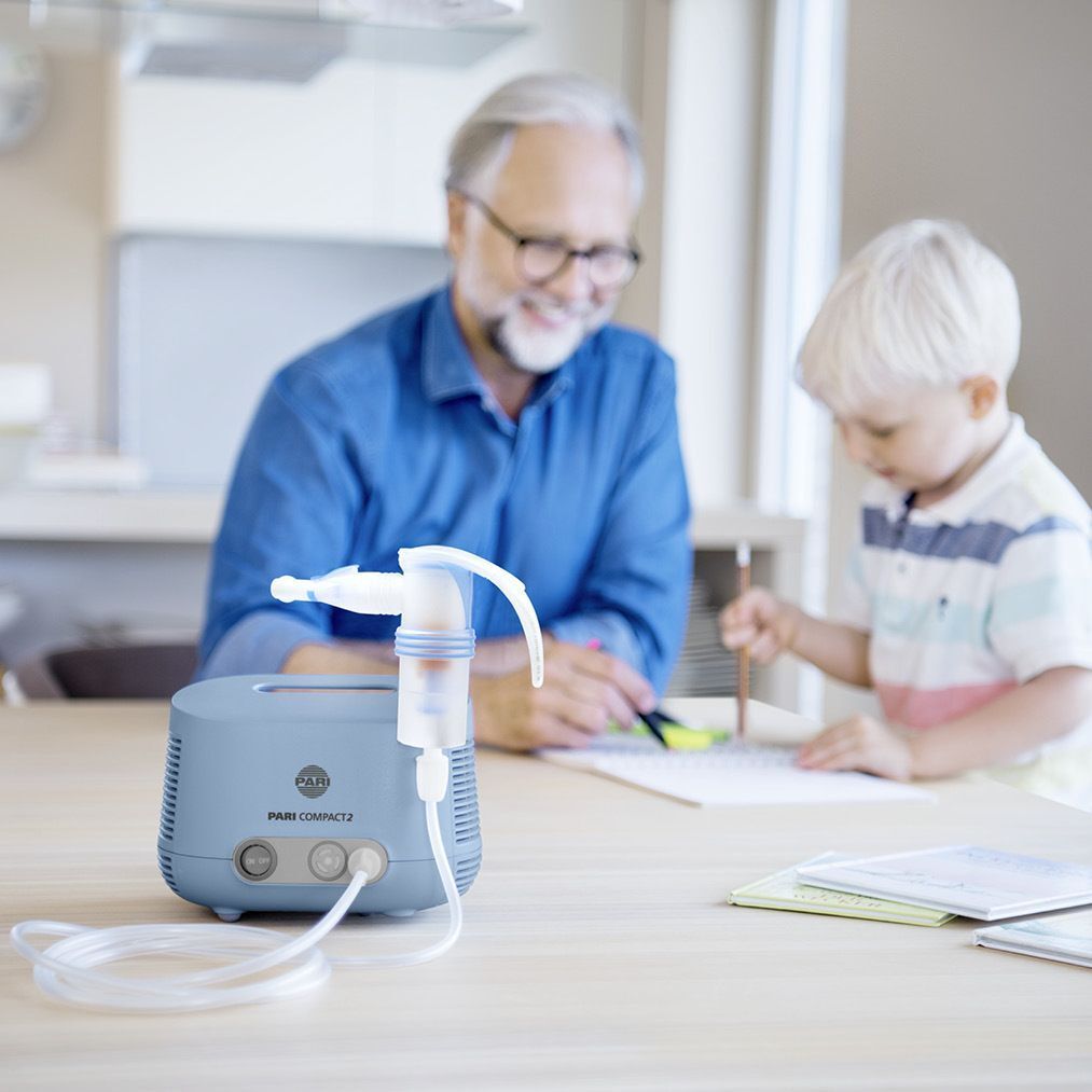 Ein Mann und ein Kind sitzen an einem Tisch. Ein PARI COMPACT 2 Inhalationsgerät steht auf dem Tisch.