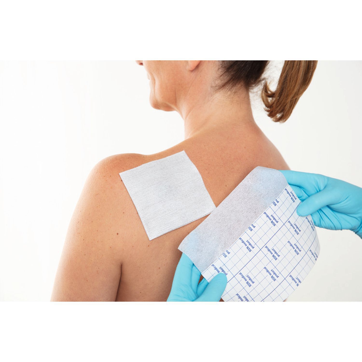 Person mit blauen Handschuhen hält Verband und Trägerpapier. Auf dem Trägerpapier ist ein Raster aufgedruckt.