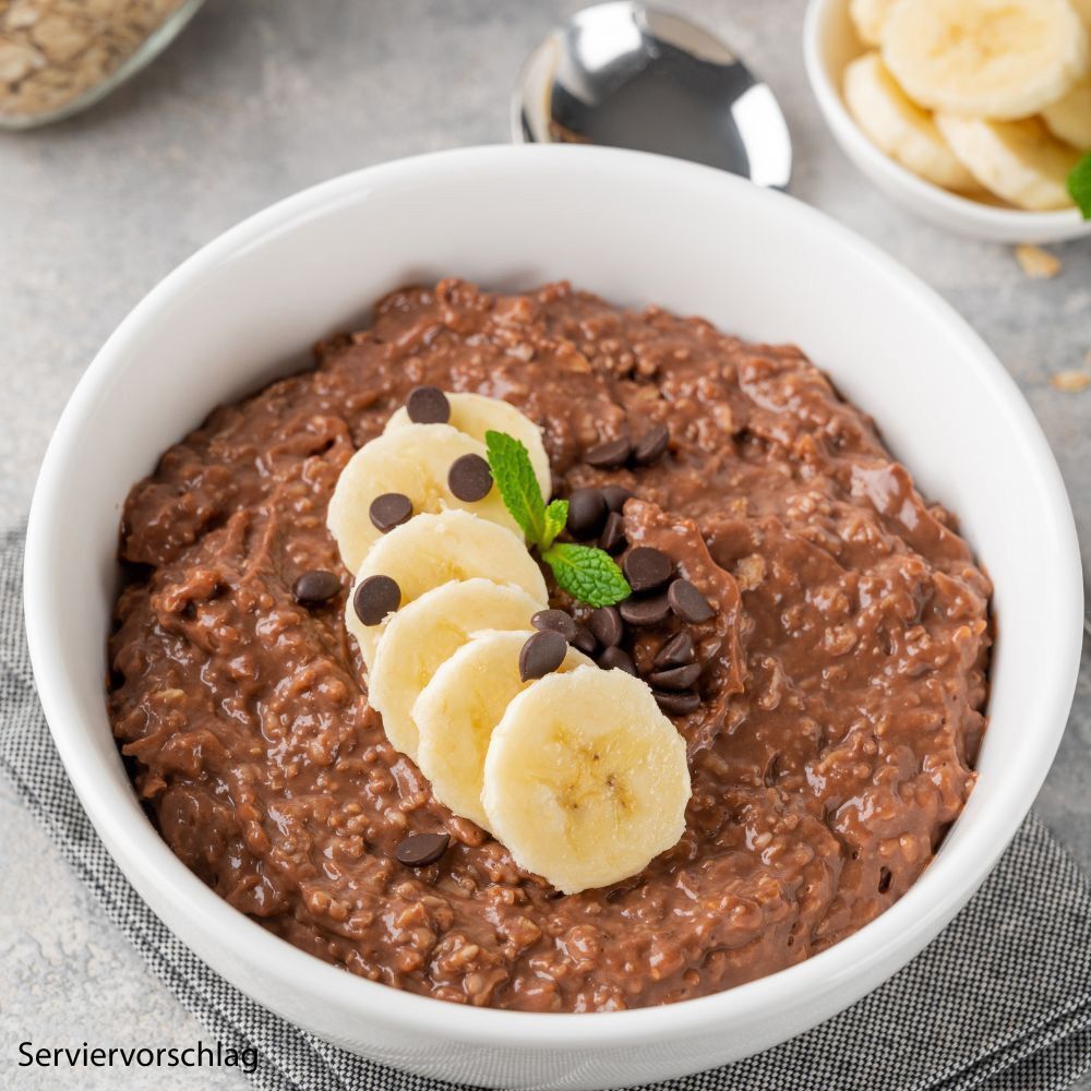 Schokoladen-Haferbrei in einer weißen Schüssel. Dekoriert mit Bananenscheiben und Schokostückchen.