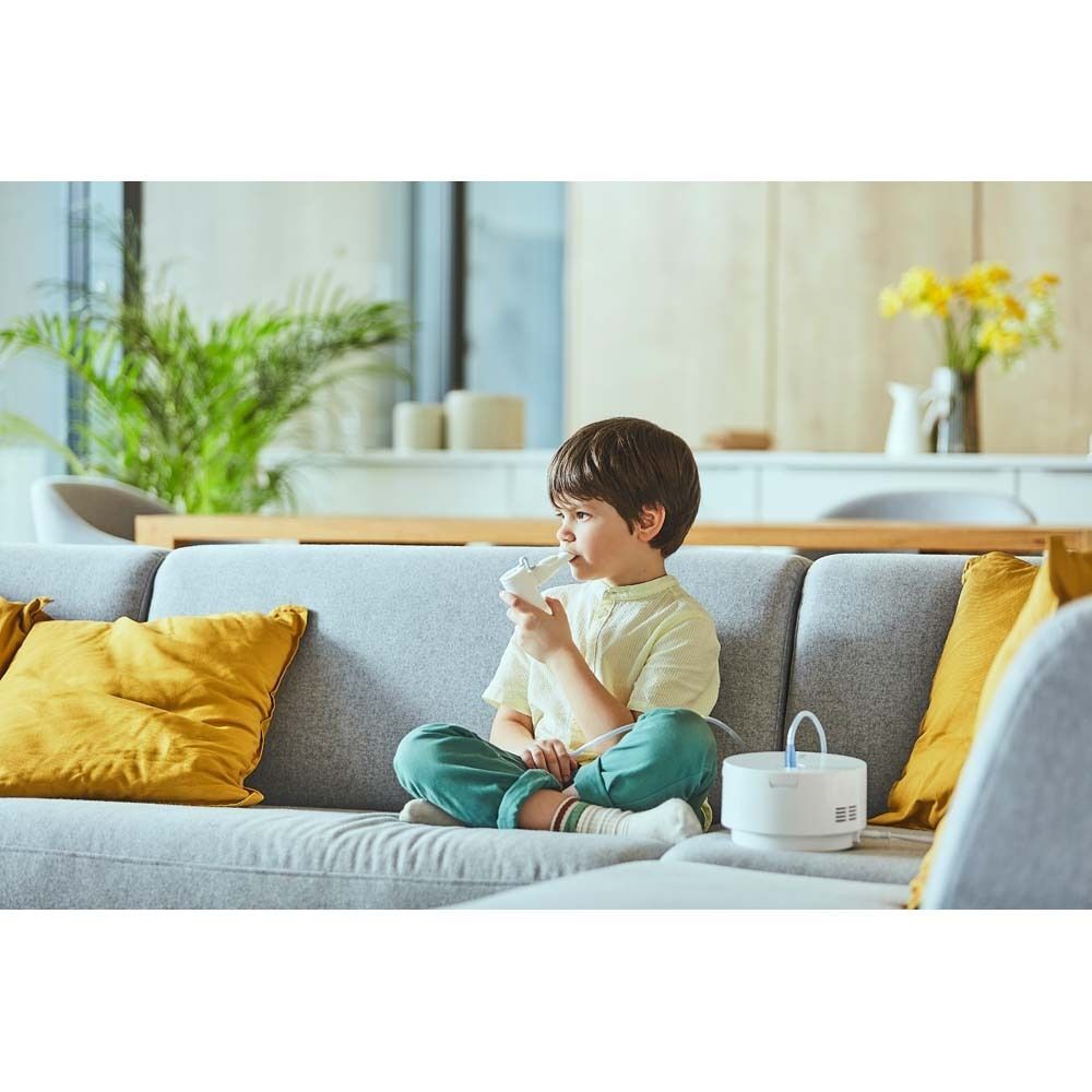 Kind benutzt ein OMRON C28P Inhaliergerät. Es sitzt auf einem Sofa. Das Gerät steht daneben.