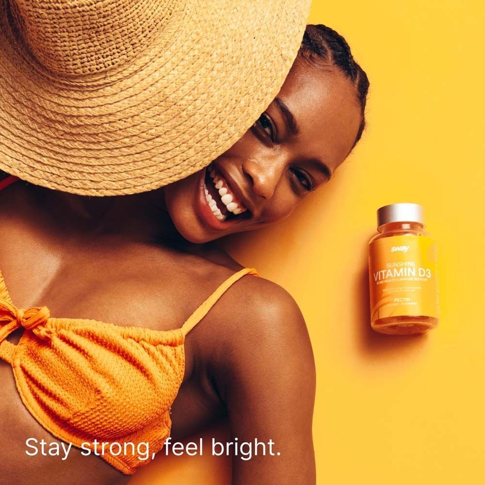 Frau mit Sonnenhut und Bikini. Flasche Vitamin D neben ihr. Gelber Hintergrund. Aufschrift: Vitamin D.