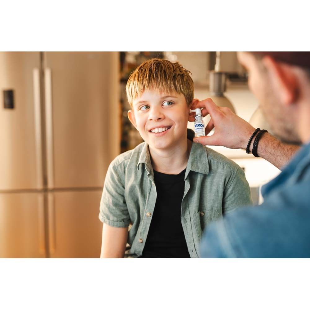 Ein Mann gibt einem lächelnden Jungen VAXOL Ohrenspray ins Ohr. Küche im Hintergrund.