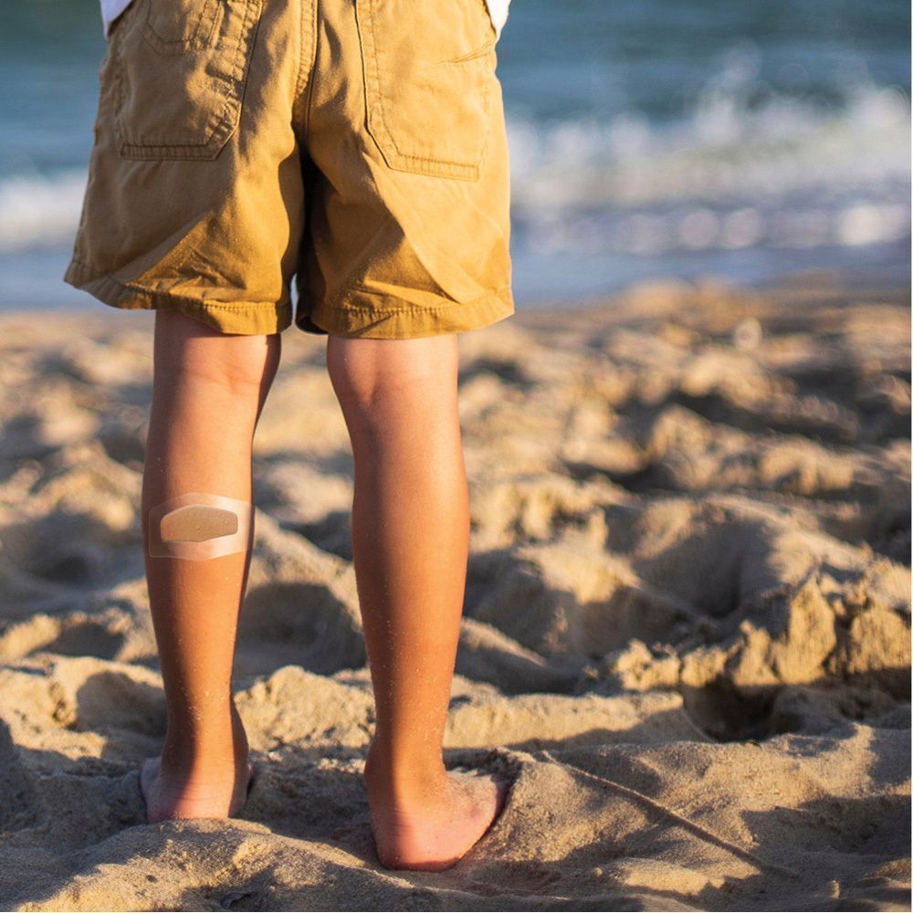 Ein Kind mit einem Pflaster am Bein. Das Pflaster ist transparent und kaum sichtbar. Strandhintergrund.