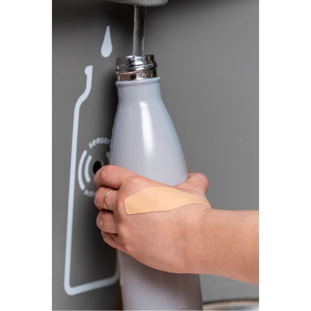 Hand mit Pflaster an einer grauen Wasserflasche. Wasser fließt aus dem Ausguss.