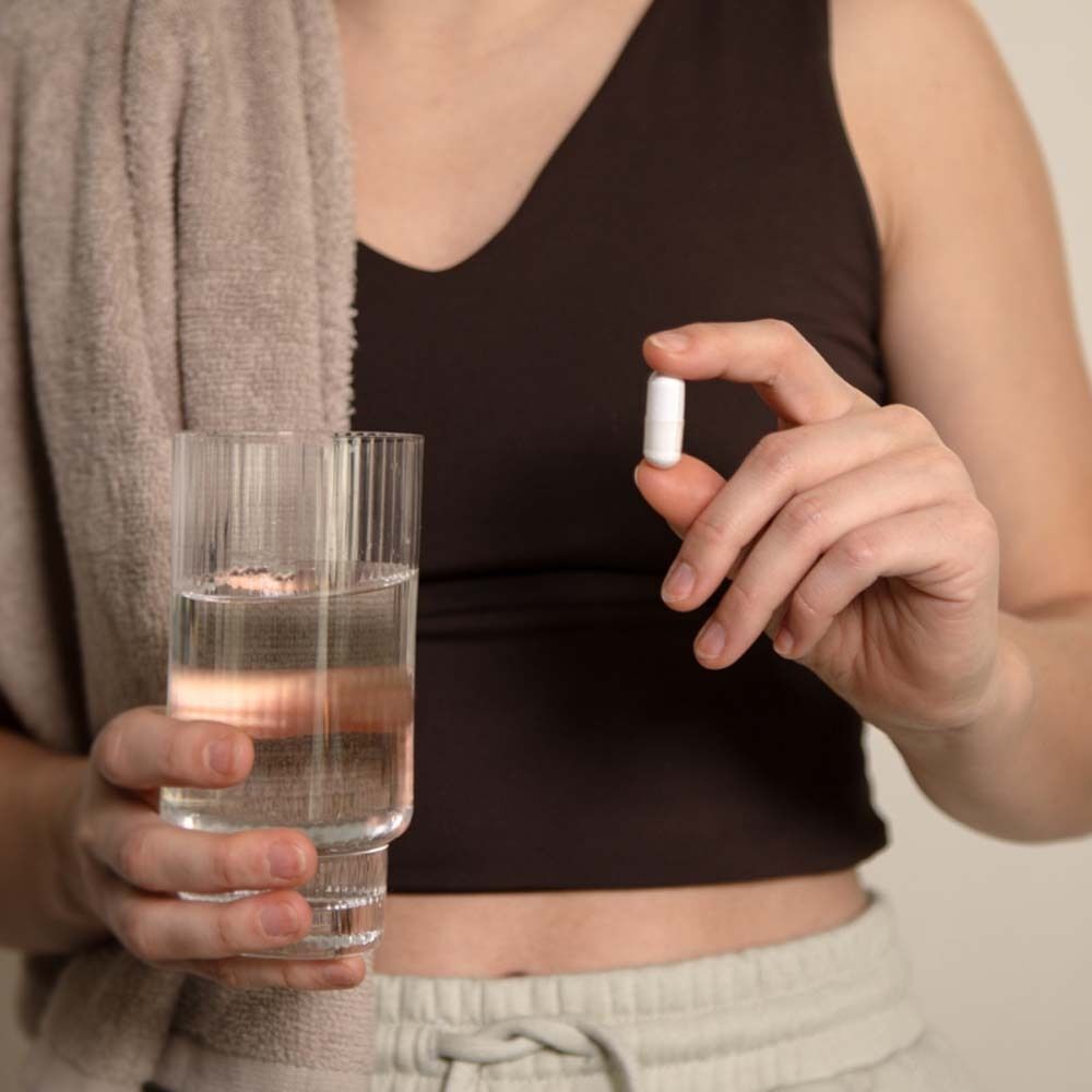 Person hält Kapsel zwischen den Fingern. Daneben Glas Wasser und Handtuch. Person trägt Top.
