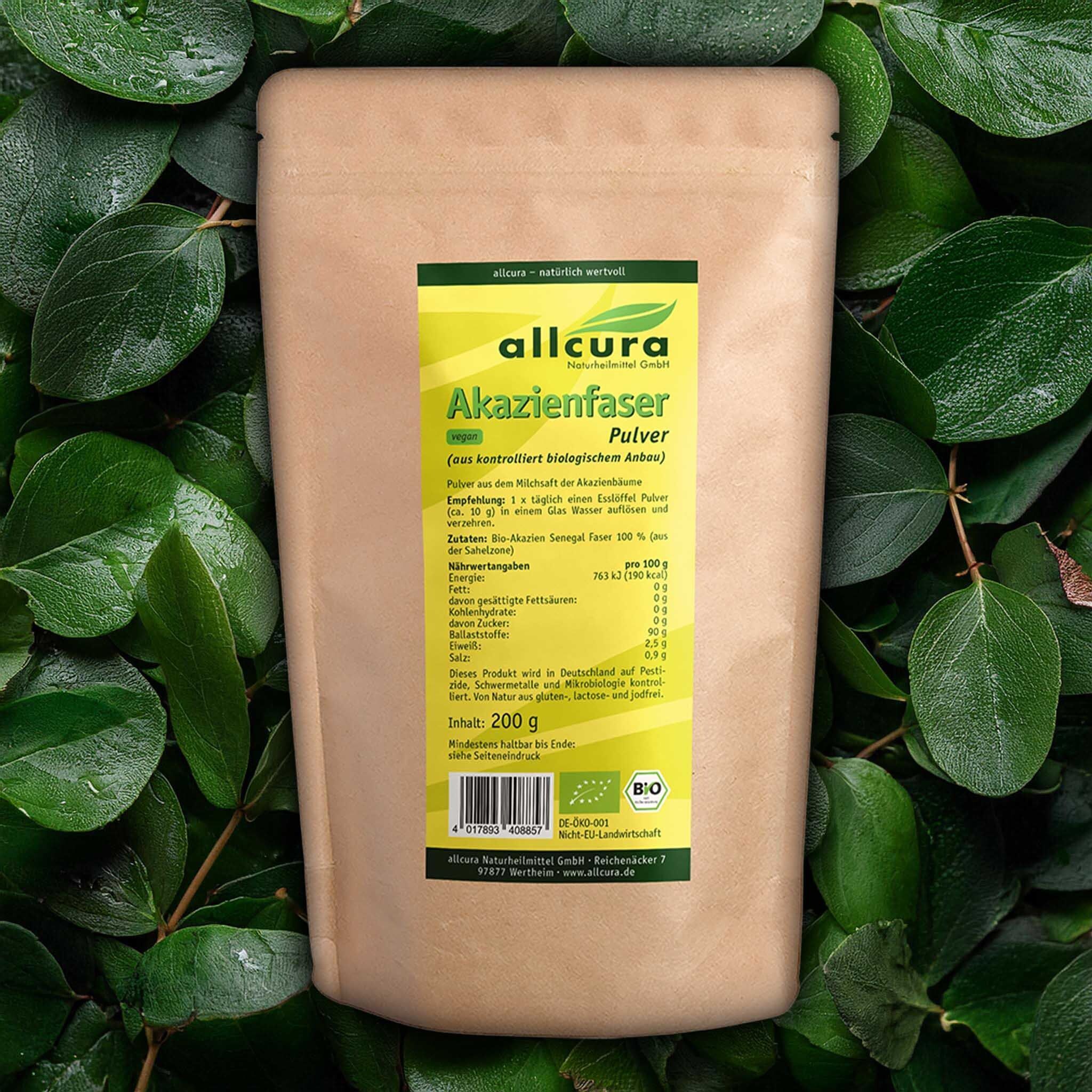 Beutel AKAZIENFASER Pulver Bio auf Blättern. Braune Papierverpackung mit gelbem Etikett. Aufschrift: allcura, Akazienfaser Pulver, Bio. 200 g. Bio-Siegel.