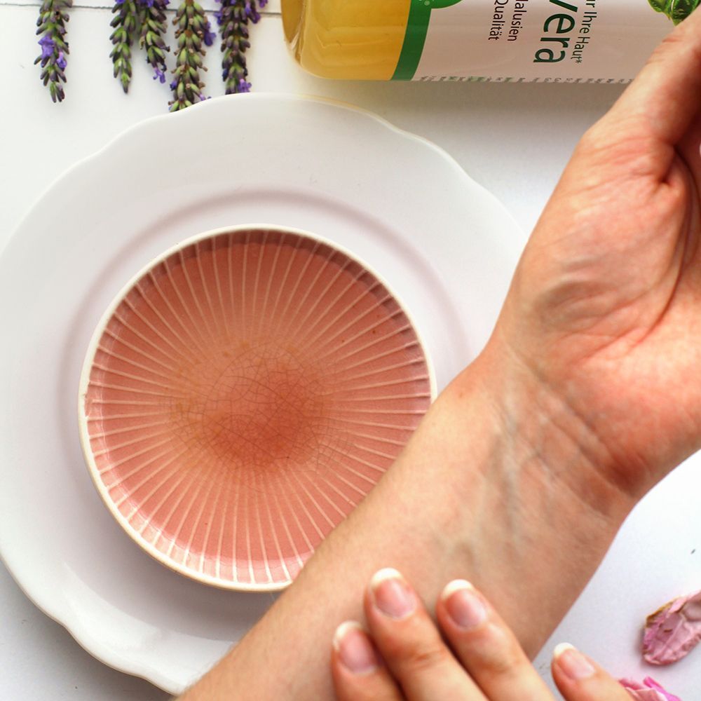 Eine Hand trägt Aloe Vera Gel auf den Unterarm auf, neben einem Teller und einer Flasche des Produkts.