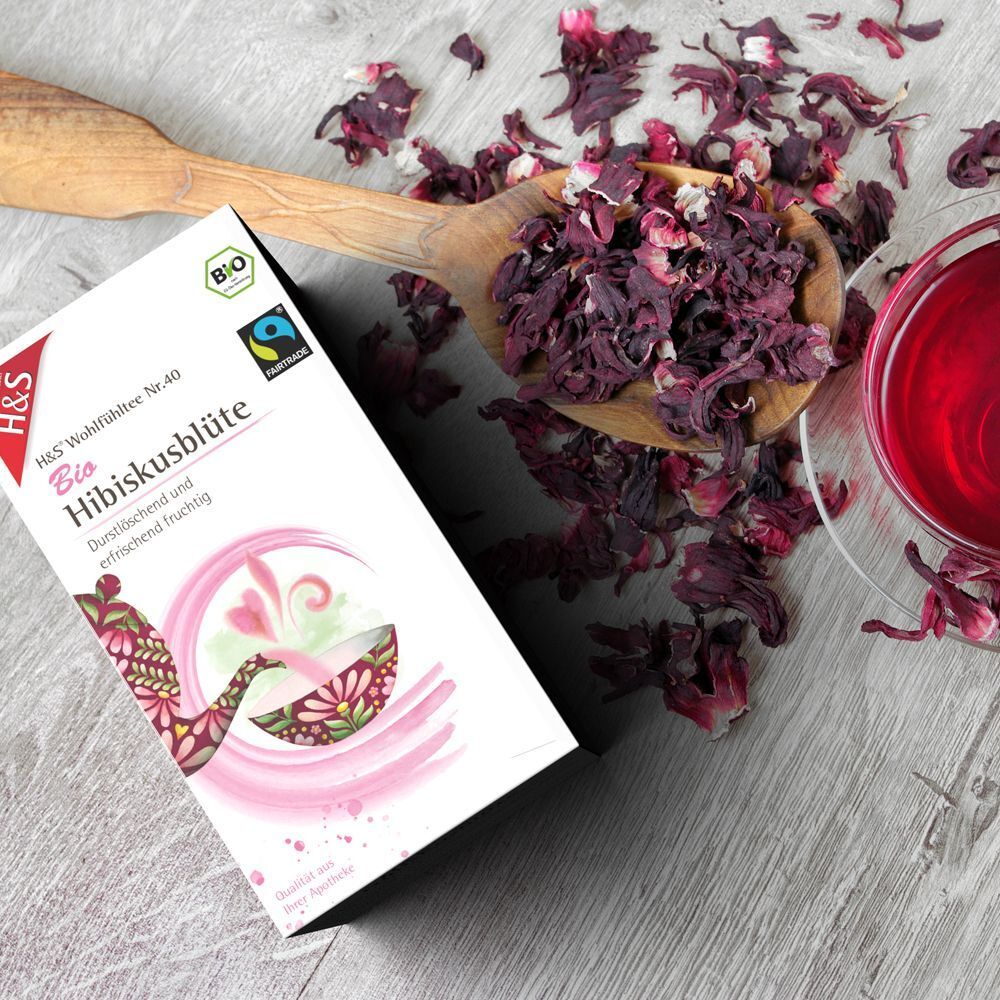 Packung H&S Bio Hibiskusblüte Filterbeutel neben Holzlöffel mit getrockneten Blüten und Glas mit rotem Tee.