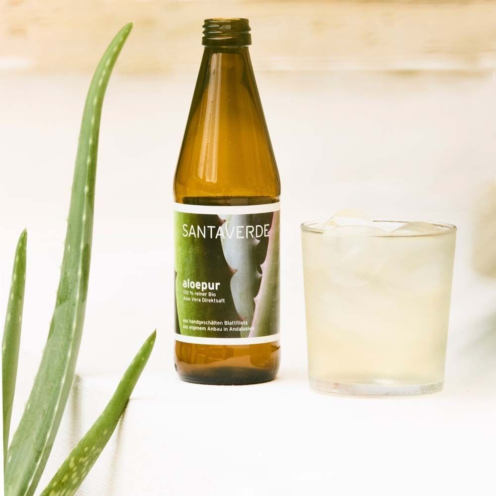 Eine Glasflasche SANTAVERDE Aloe Vera Saft neben einem Glas mit Eiswürfeln und einem Aloe Vera Blatt.