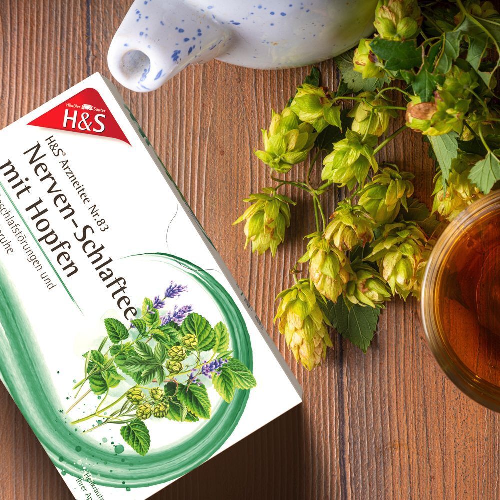 Packung H&S Nerven-Schlaftee mit Hopfen auf Holztisch, neben Hopfen und einer Teetasse.