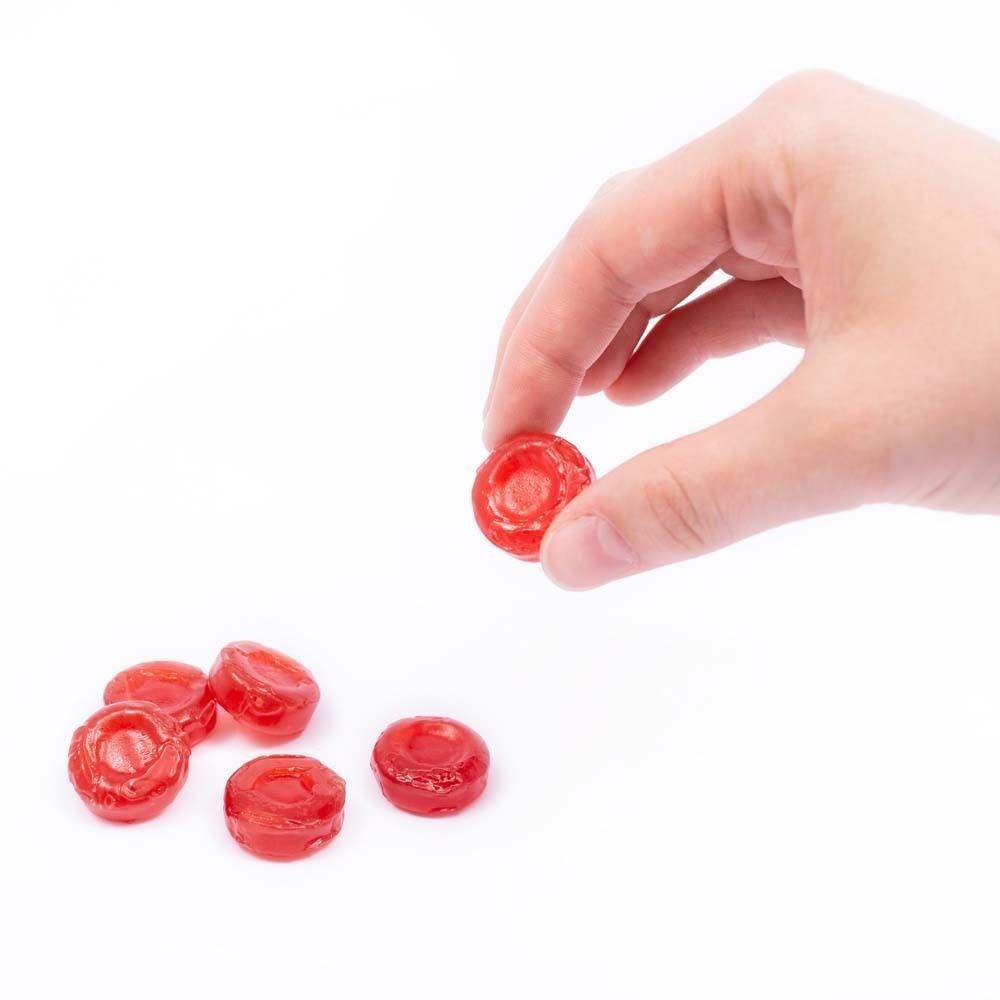 Hand hält eine rote Hustenbonbon. Mehrere weitere Bonbons liegen daneben. Runde Form, leicht transparent.
