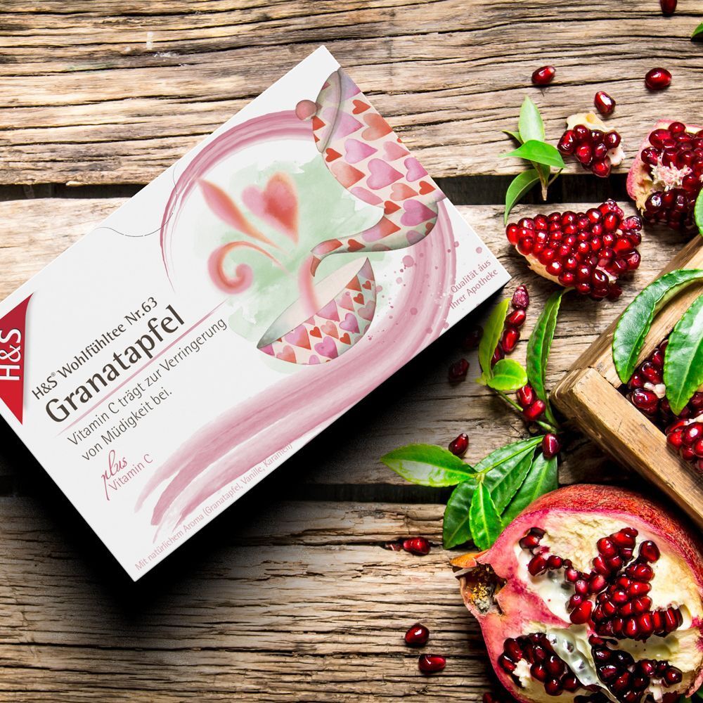 H&S Granatapfel Teebeutel-Packung neben Granatapfel-Hälften und -Kernen auf Holz. Produktname und Logo sichtbar. Rosa Design.