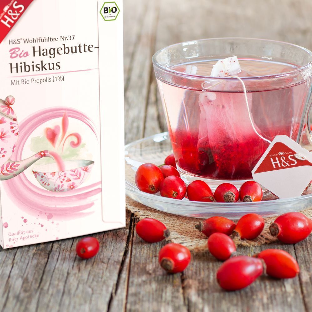 Glas Tee mit Teebeutel. Daneben Hagebutten und eine Packung Tee. Aufdruck: H&S Bio Hagebutte-Hibiskus.