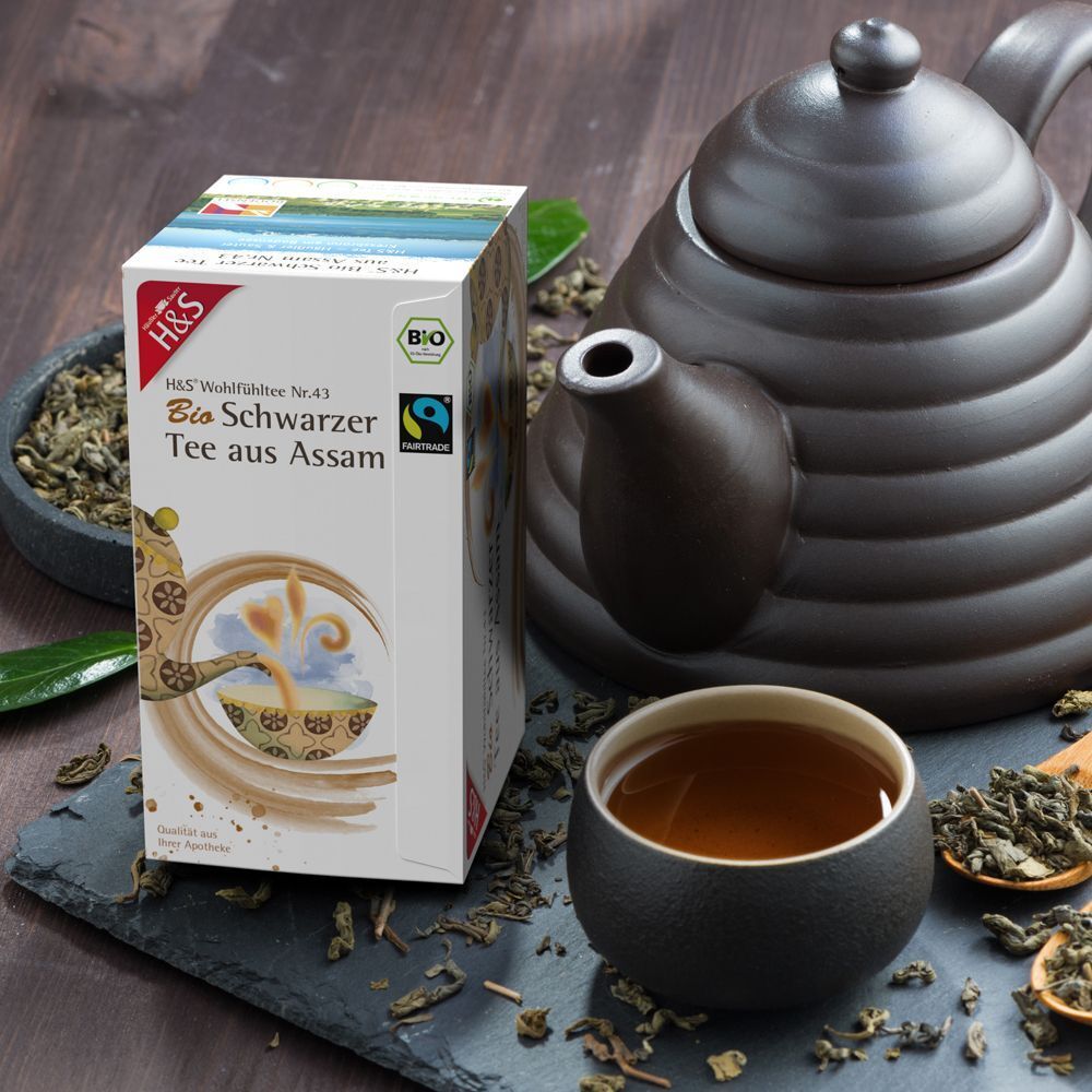 H&S Bio Schwarzer Tee aus Assam Packung neben Teekanne und Tasse. Teebeutel und loser Tee auf Schieferplatte.