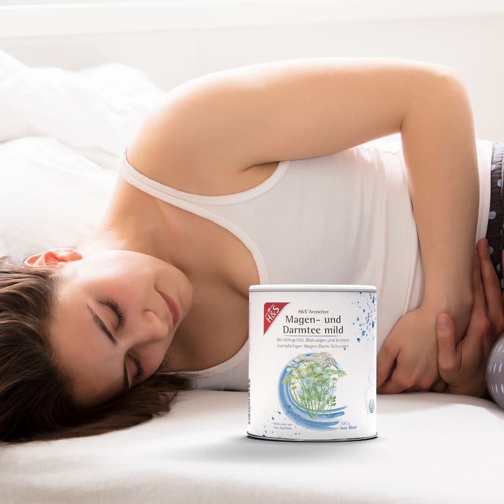 Eine Frau liegt mit Bauchschmerzen im Bett, daneben steht eine Dose H&S Magen- und Darmtee mild.