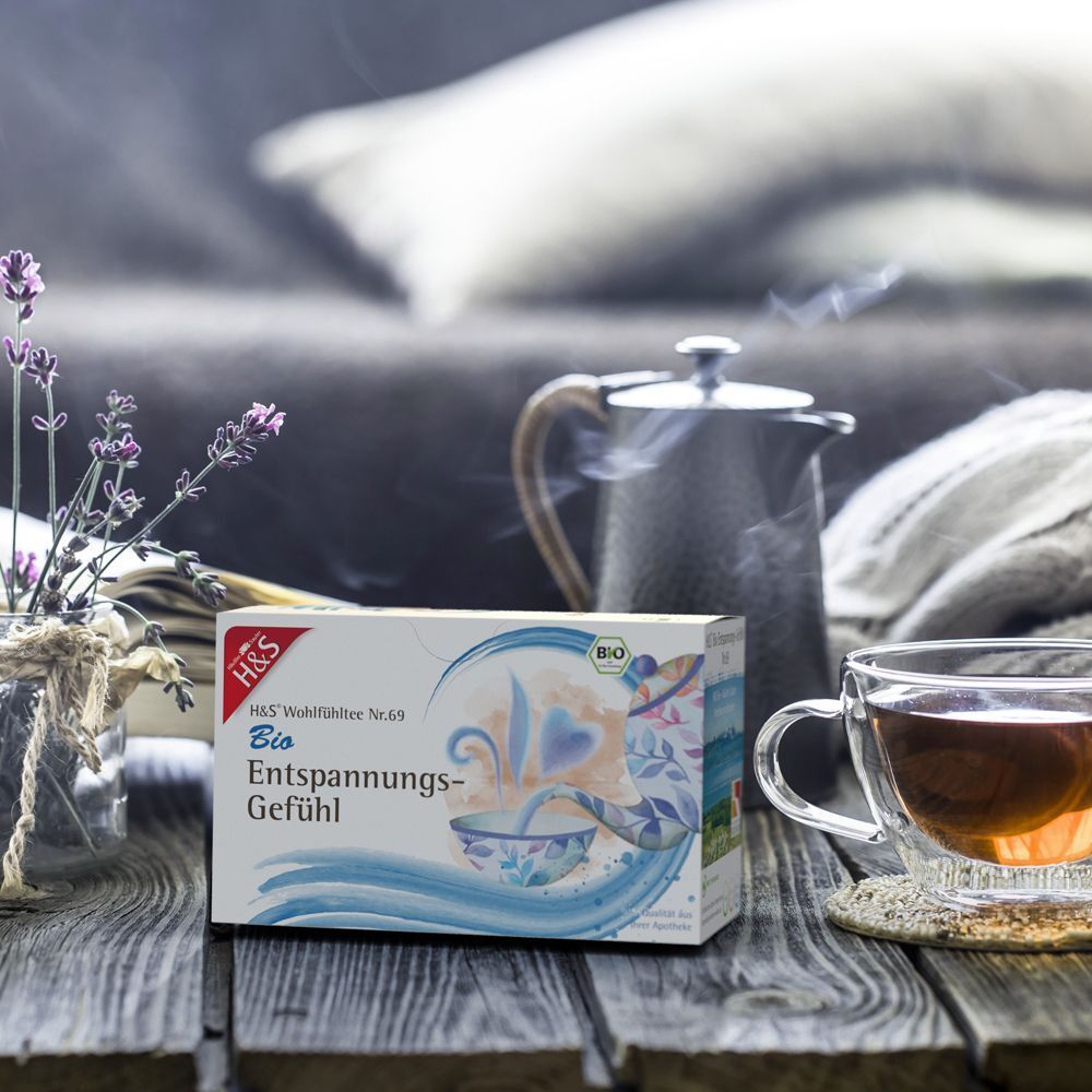 H&S Bio Entspannungs-Gefühl Tee-Packung neben Teekanne und Tasse. Auf Holztisch. Lavendelblüten.