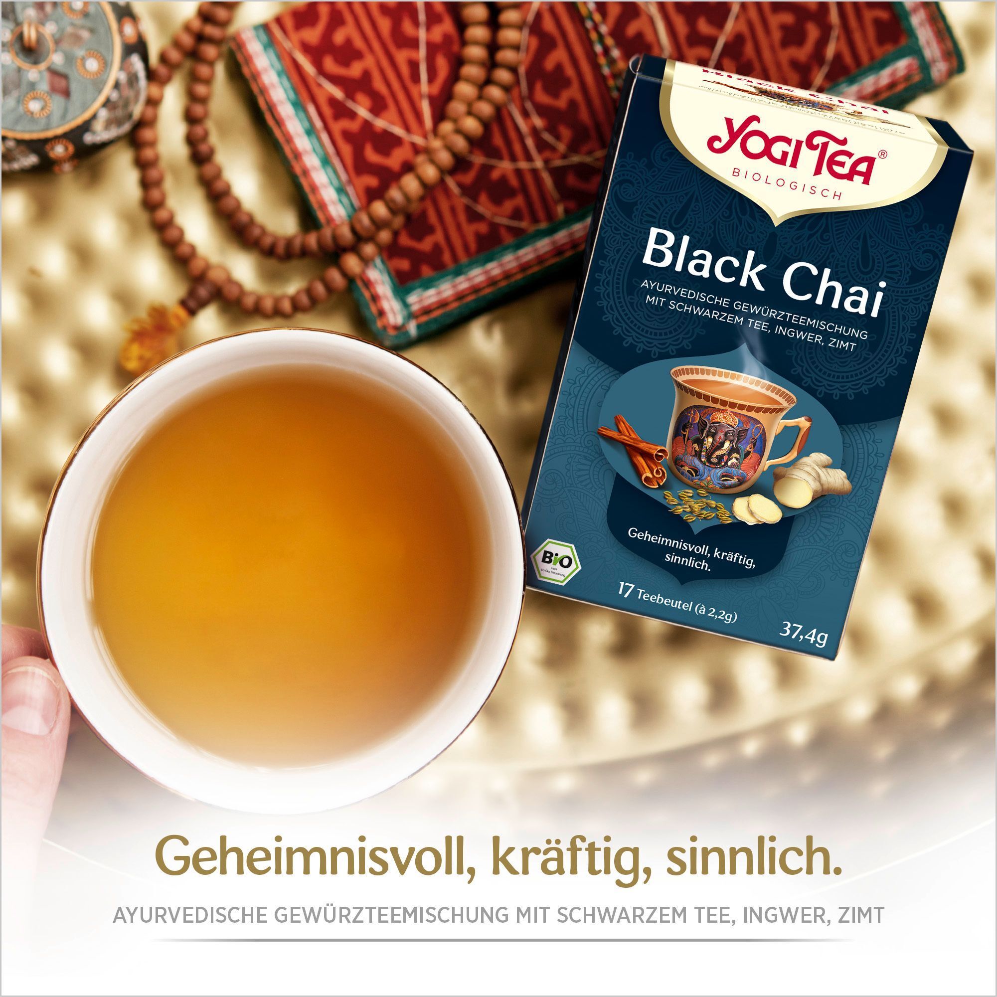 YOGI TEA Black Chai Bio Filterbeutel neben einer Tasse Tee. Aufschrift: Black Chai, ayurvedische Gewürzmischung mit schwarzem Tee, Ingwer, Zimt. 17 Teebeutel.