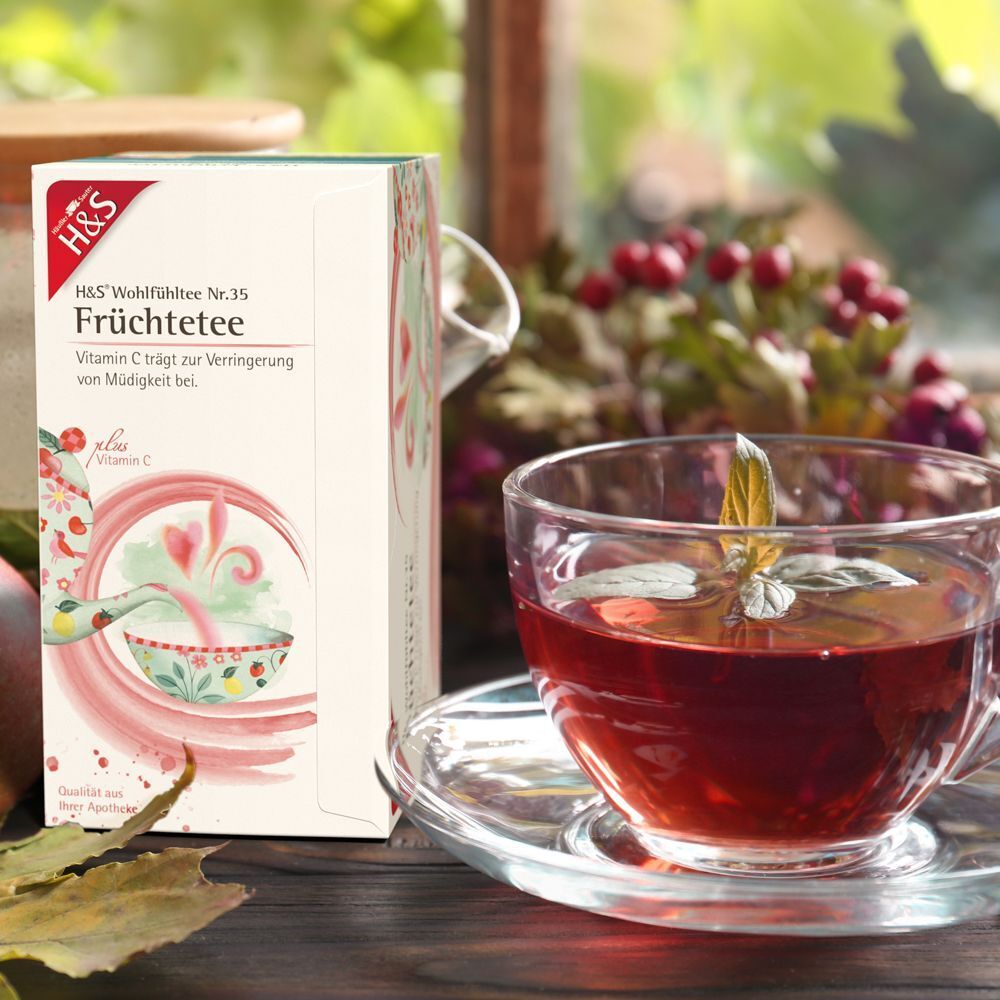 H&S Früchtetee-Packung neben einer Teetasse. Die Tasse enthält roten Tee. Im Hintergrund ein Fenster und Pflanzen.