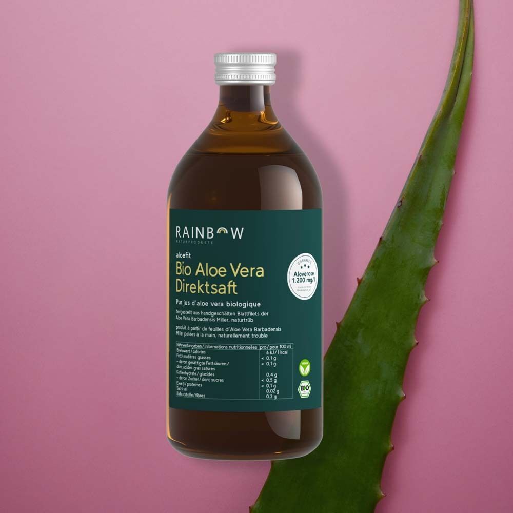 Eine Flasche Rainbow Aloefit Bio Aloe Vera Direktsaft neben einem Aloe Vera Blatt auf rosa Hintergrund. Das Etikett ist lesbar.