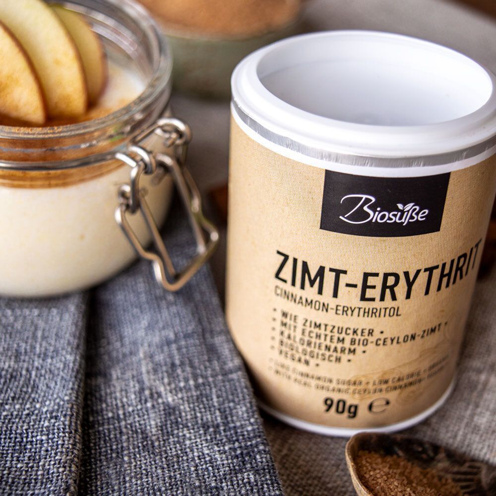 Dose Biosüße Zimt-Erythrit neben Glas mit Dessert und Apfelscheiben. Braune Dose, schwarzes Etikett. Text: Zimt-Erythrit, 90g.