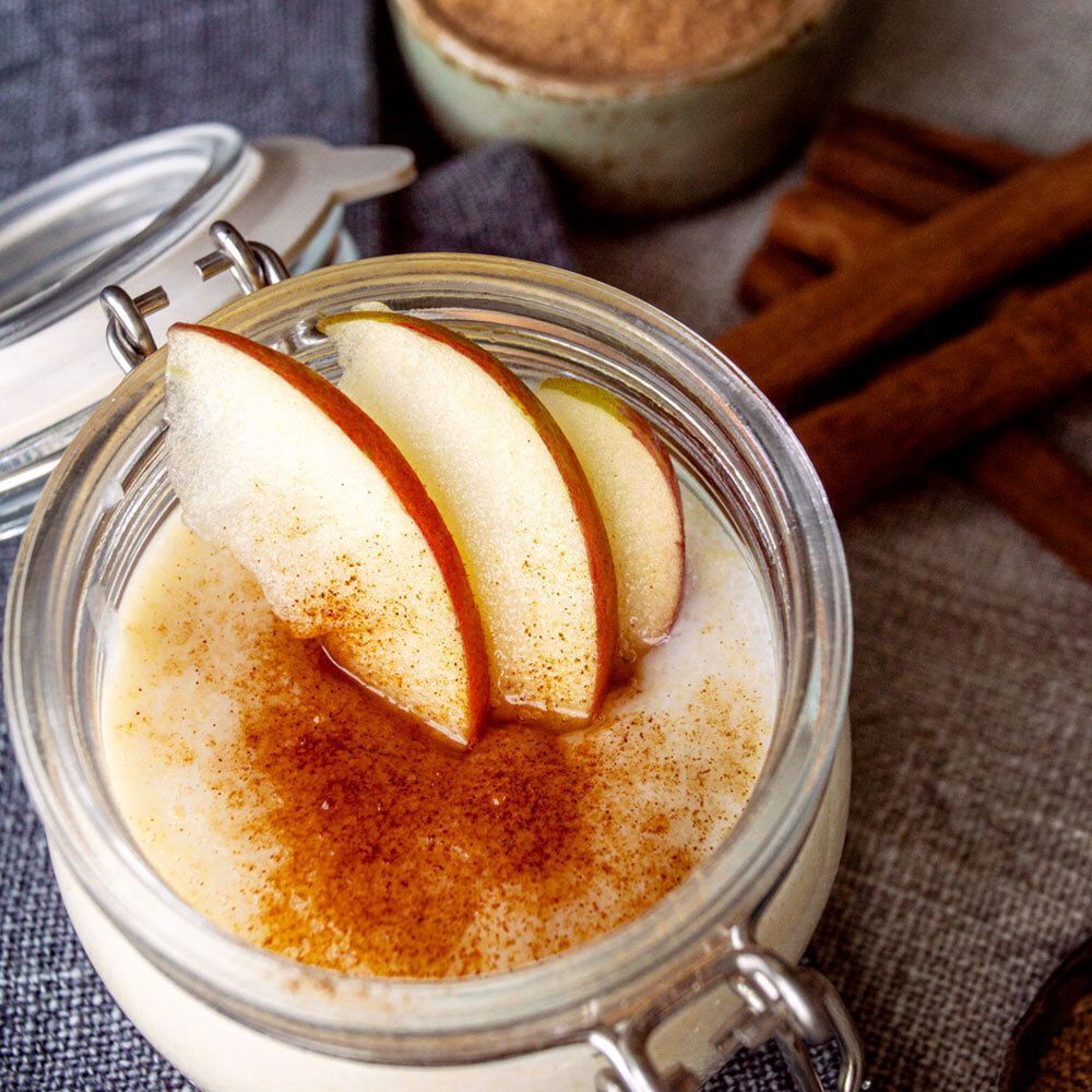 Glas mit Dessert, bestreut mit Zimt, und Apfelscheiben. Auf grauem Stoff. Im Hintergrund: Zimtstangen und Schale mit Zimt.