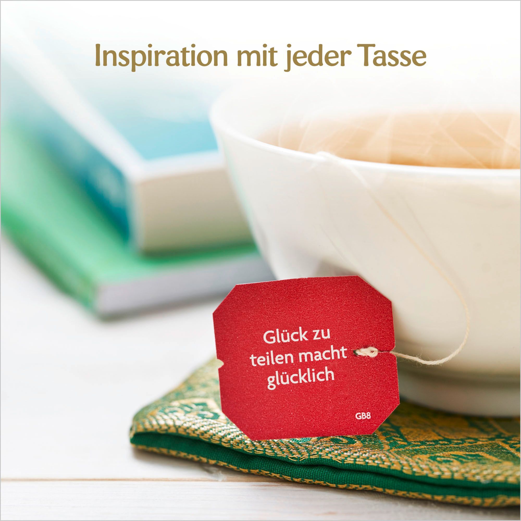 Tasse Tee mit rotem Teebeutel. Aufschrift: Glück zu teilen macht glücklich. Bücher im Hintergrund. Grünes Kissen.