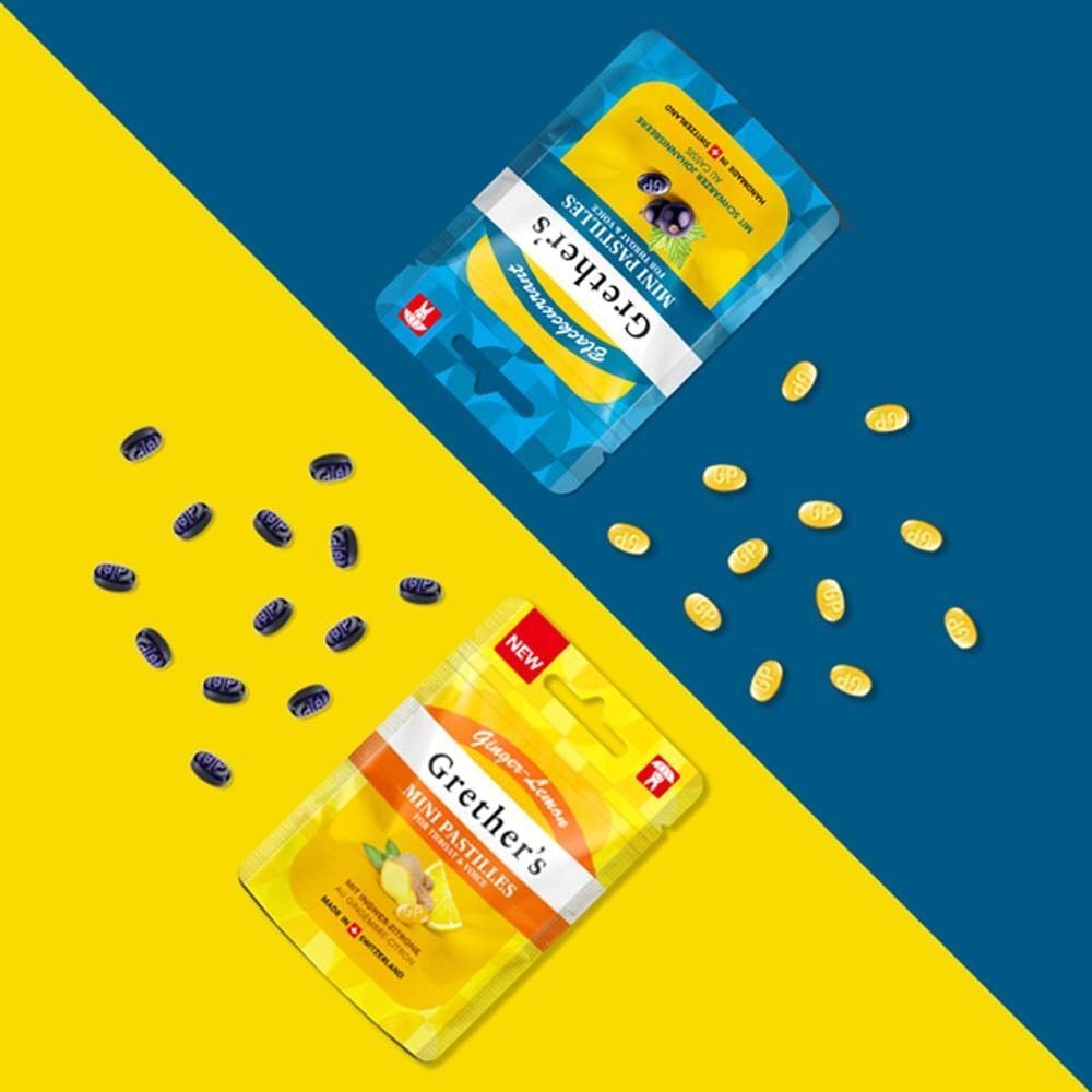 Zwei Beutel Grether's Mini Pastilles, einer mit schwarzen Johannisbeeren, einer mit Ingwer-Zitrone. Pastillen verstreut. Hintergrund gelb/blau geteilt.