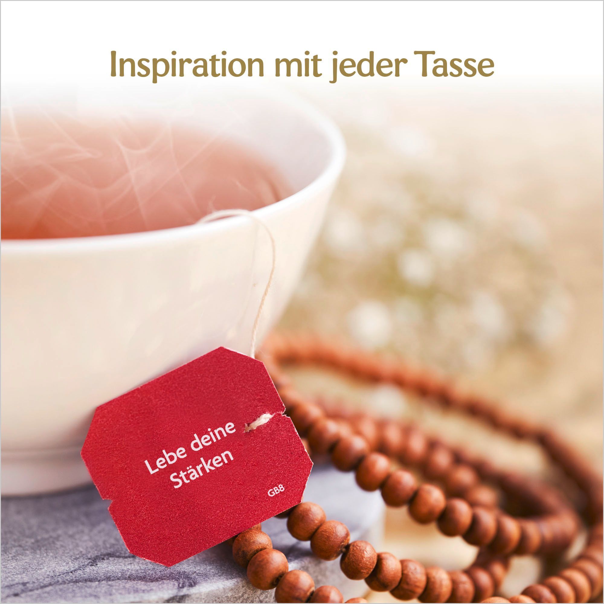 Tasse Tee mit rotem Teebeutel. Text: Lebe deine Stärken. Perlenkette. Hintergrund unscharf.