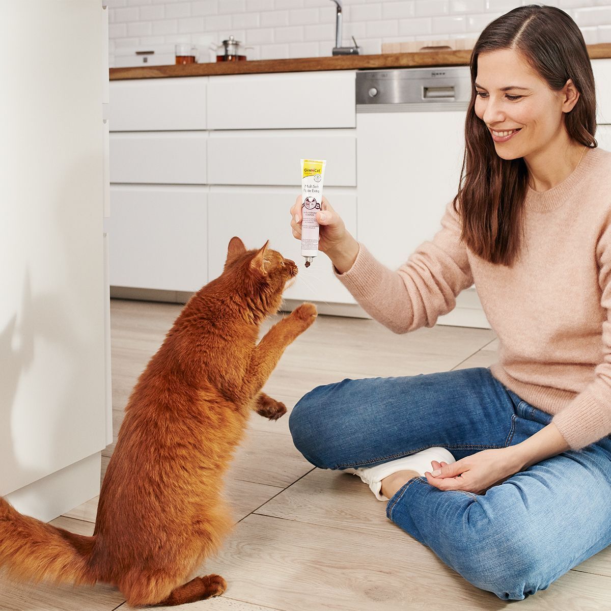 Frau und Katze. Frau hält Tube 'Malt-Soft Paste Extra' in der Hand. Katze springt. Küche im Hintergrund.