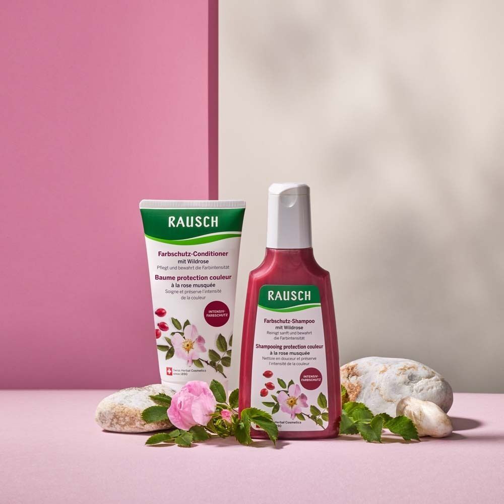 Flasche RAUSCH Farbschutz-Shampoo und Tube RAUSCH Farbschutz-Conditioner mit Wildrose, dekoriert mit Steinen und Rosen.