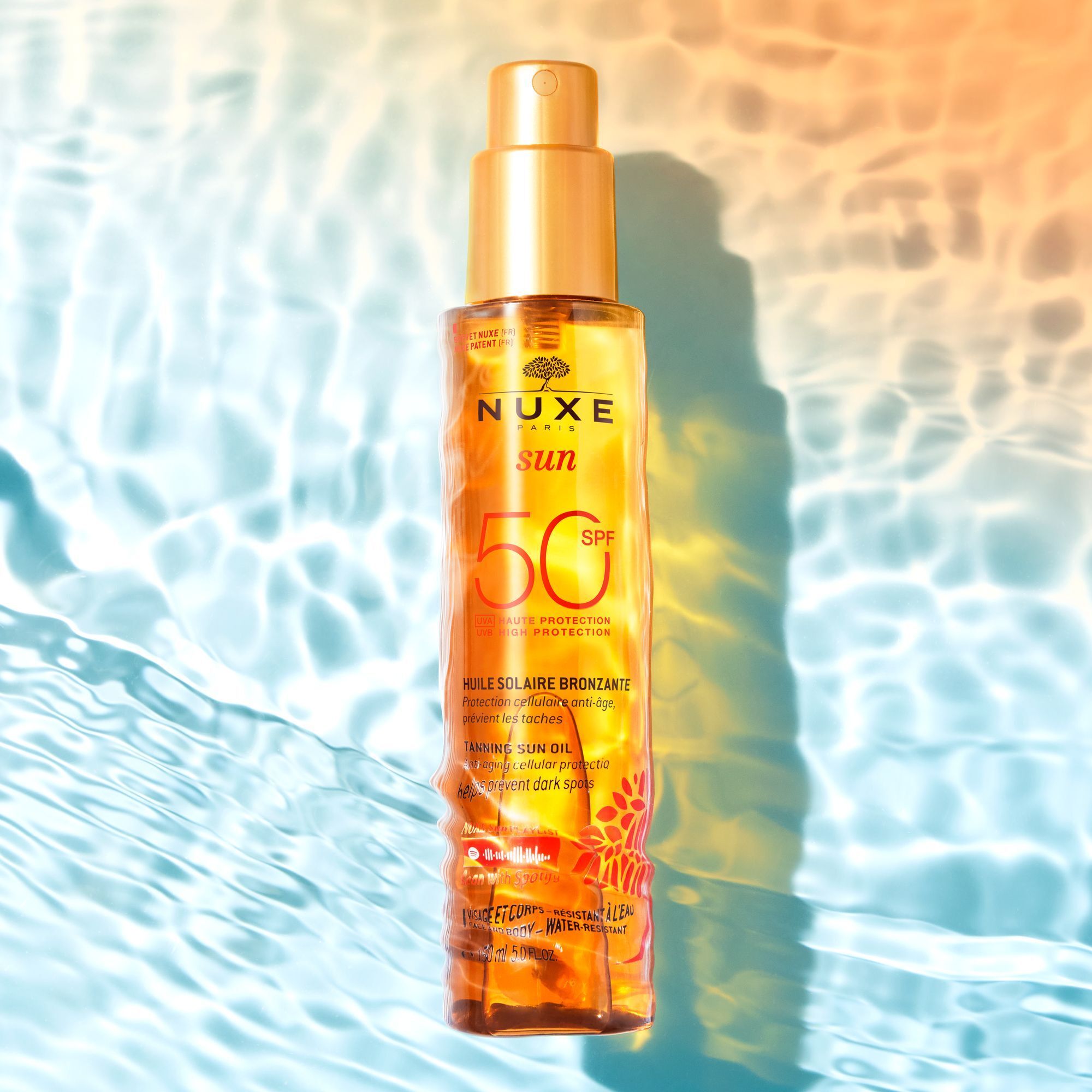 Eine Flasche Nuxe Sun Sonnenöl LSF 50 mit Pumpspray-Applikator, liegend auf einer blauen, welligen Oberfläche.