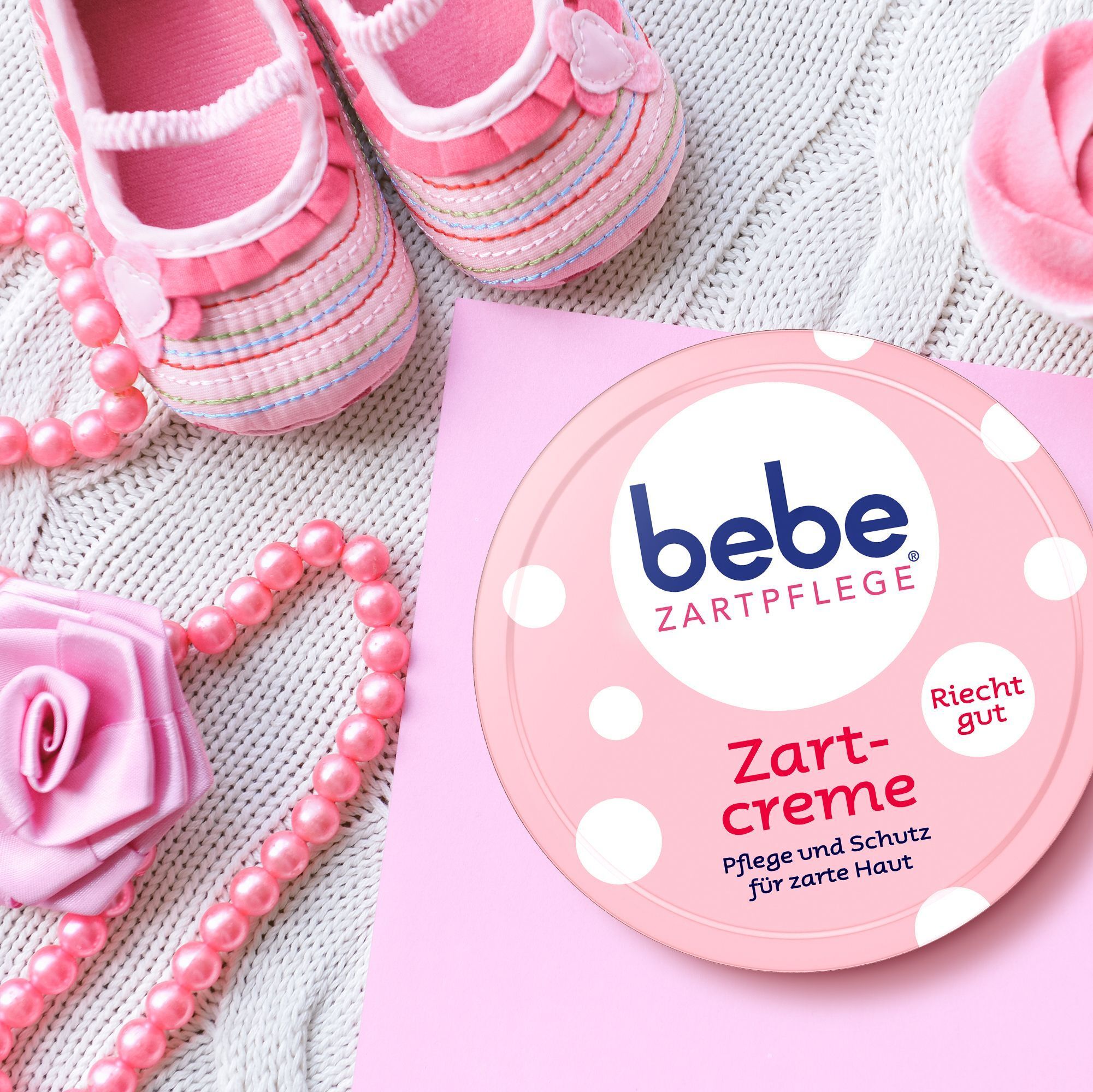 Bebe Zartcreme Dose neben Babyschuhen, Perlenkette und einer Rosenblüte.