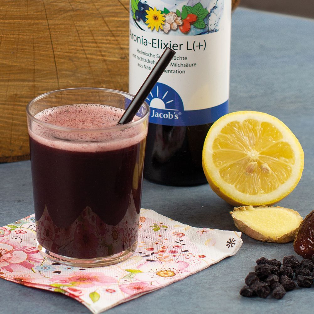 Ein Glas Aronia-Elixier mit Strohhalm, daneben Zitrone, Ingwer und Aroniabeeren.