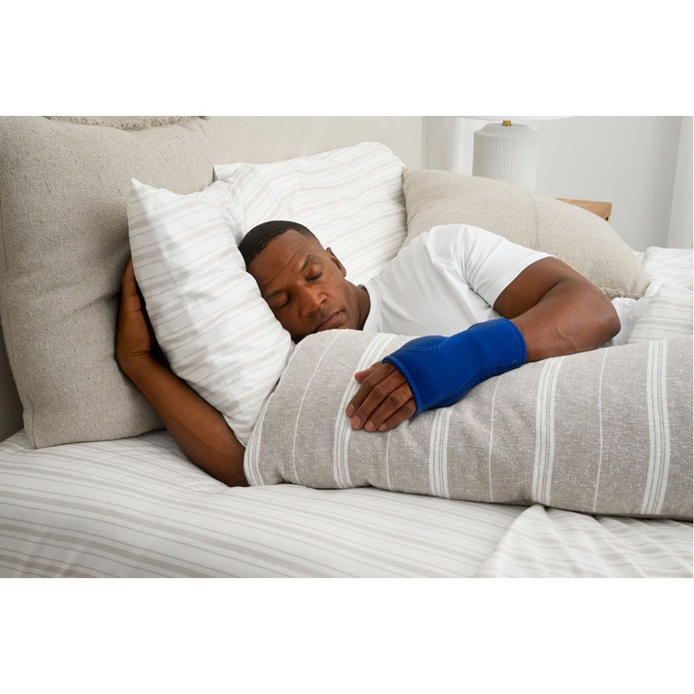 Person schläft mit blauer Handgelenkbandage am Arm. Liegt auf dem Bett. Kissen und Decke im Hintergrund.