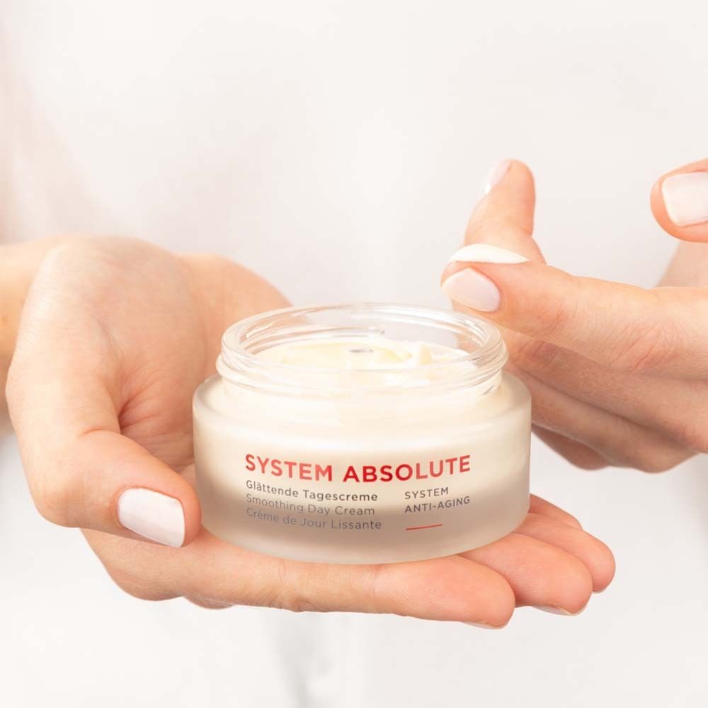Hände halten einen Cremetiegel. Aufschrift: SYSTEM ABSOLUTE, Glättende Tagescreme, Smoothing Day Cream, Crème de Jour Lissante.