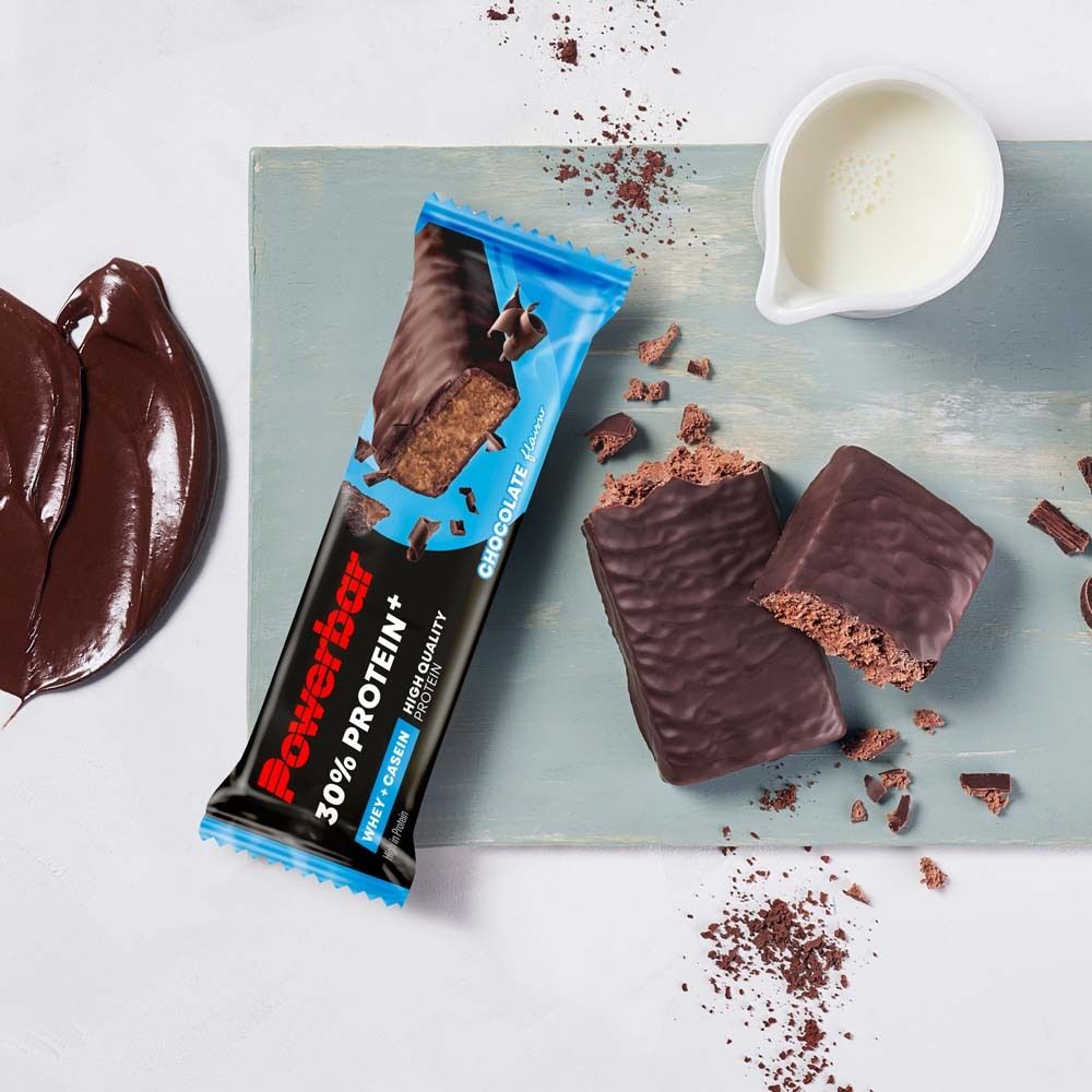 Proteinriegel mit Schokoladenüberzug. Auf einem Holzbrett. Daneben Milch und Schokoladenstückchen.