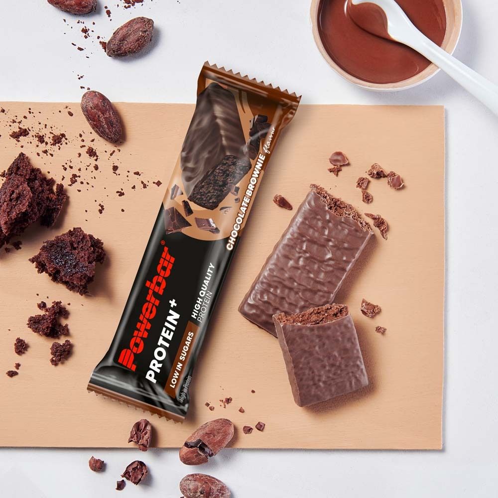 Powerbar PROTEIN+ LOW SUGAR CHOCOLATE BROWNIE. Riegel und Stücke auf Holzbrett. Kakaobohnen und Schokoladensoße.