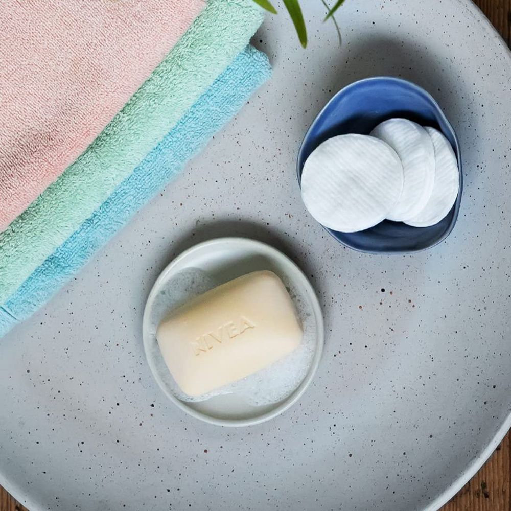 NIVEA Creme Soft Seife auf einem Seifenhalter, umgeben von Schaum. Daneben liegen Wattepads und Handtücher.