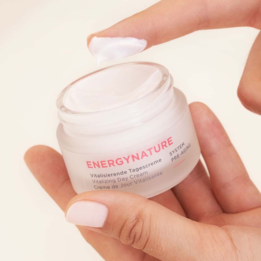 Hand hält Tiegel mit Creme. Aufschrift ENERGYNATURE, Vitalisierende Tagescreme. Ein Finger berührt Creme.