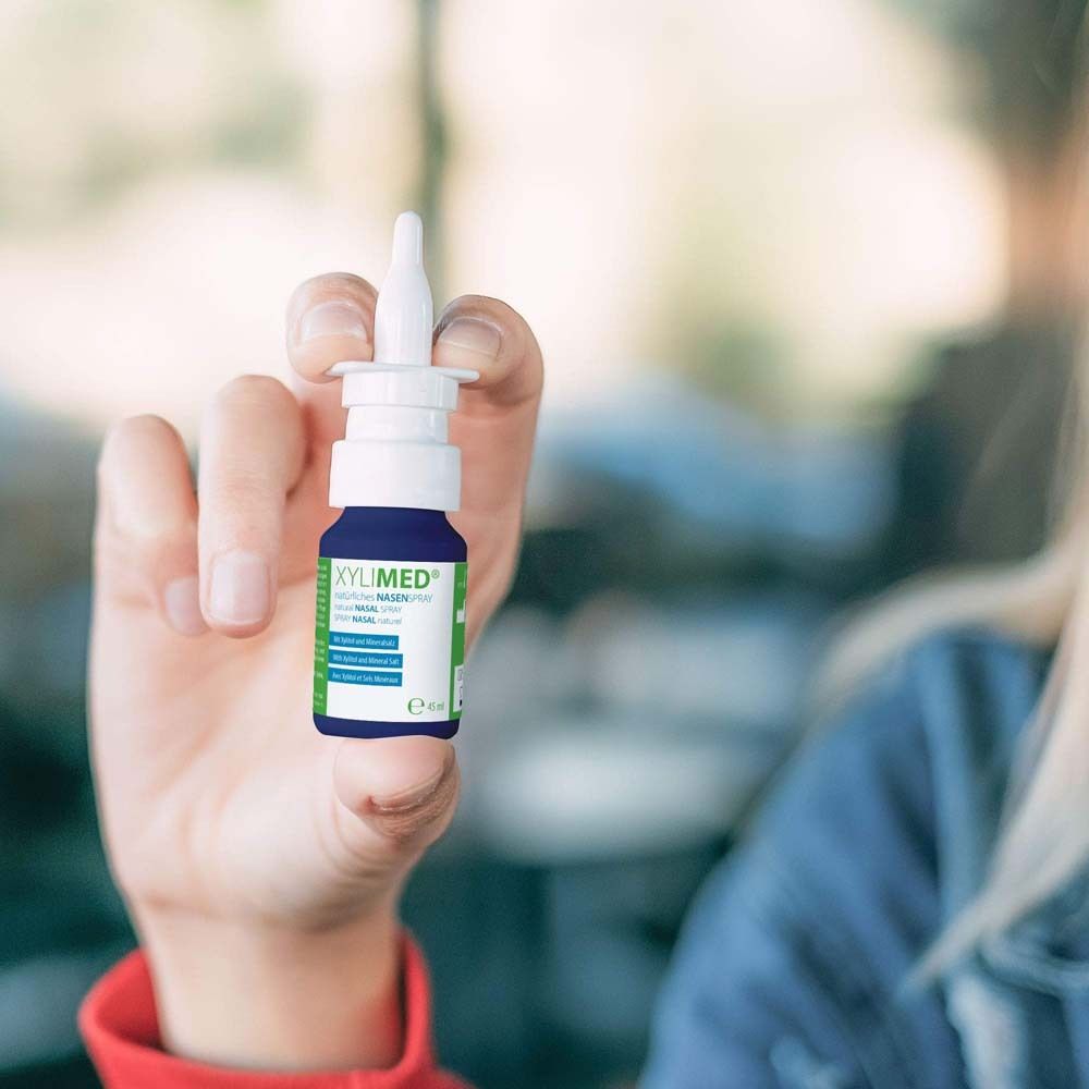 Person hält eine blaue Nasenspray-Flasche. XYLIMED® Nasenspray, weißer Sprühkopf, grüne und weiße Etiketten.