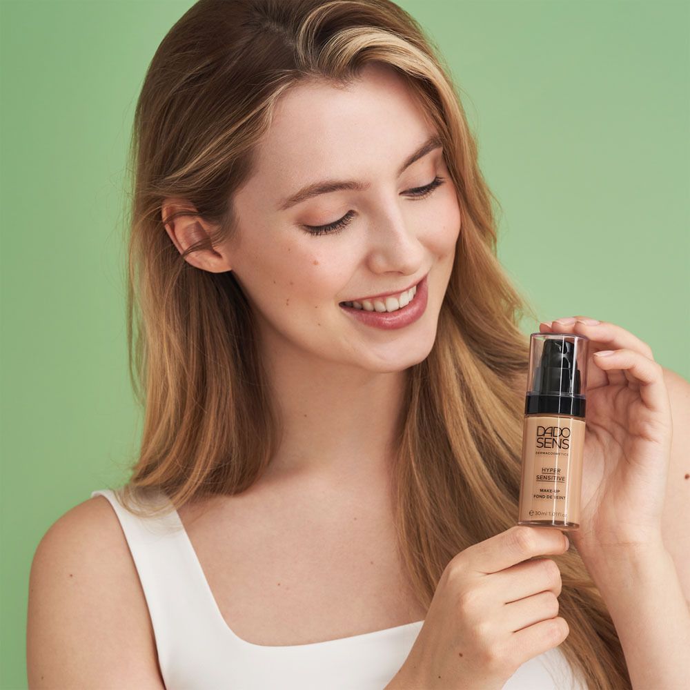 Frau hält Dado Sens Make-up-Fläschchen. Produktname und "Hypersensitive" sichtbar. Grüner Hintergrund.