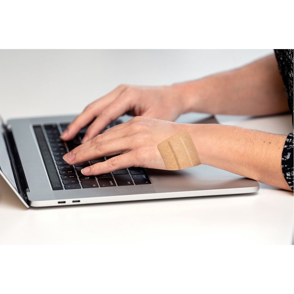 Hände tippen auf Laptop-Tastatur. Pflaster am Handgelenk. Weißer Tisch.