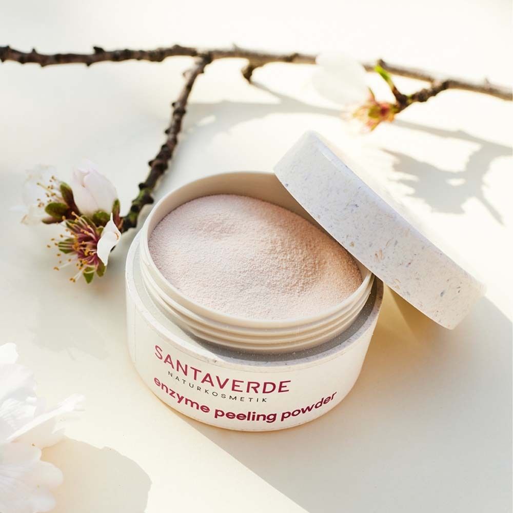 Geöffnete Dose mit SANTAVERDE enzyme peeling powder. Rosa Pulver sichtbar. Blütenzweig im Hintergrund.