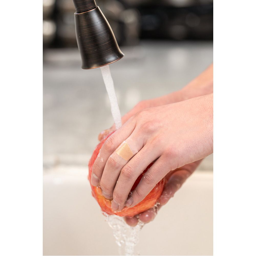 Person mit Pflaster am Finger. Person wäscht Apfel unter fließendem Wasser. Pflaster ist transparent.