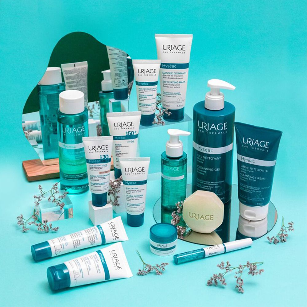 Verschiedene Uriage-Produkte auf blauem Hintergrund. Flaschen, Tuben, Tiegel. Text: Uriage, Hyséac, 100ml, 3.4 fl.oz. Spiegel und Dekoration.