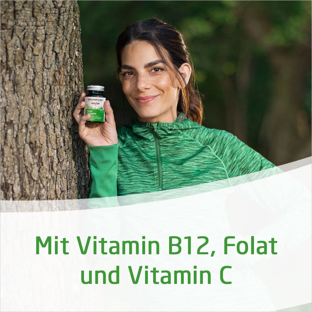 Eine Frau hält eine Flasche vor einem Baum. Sie trägt grüne Kleidung.