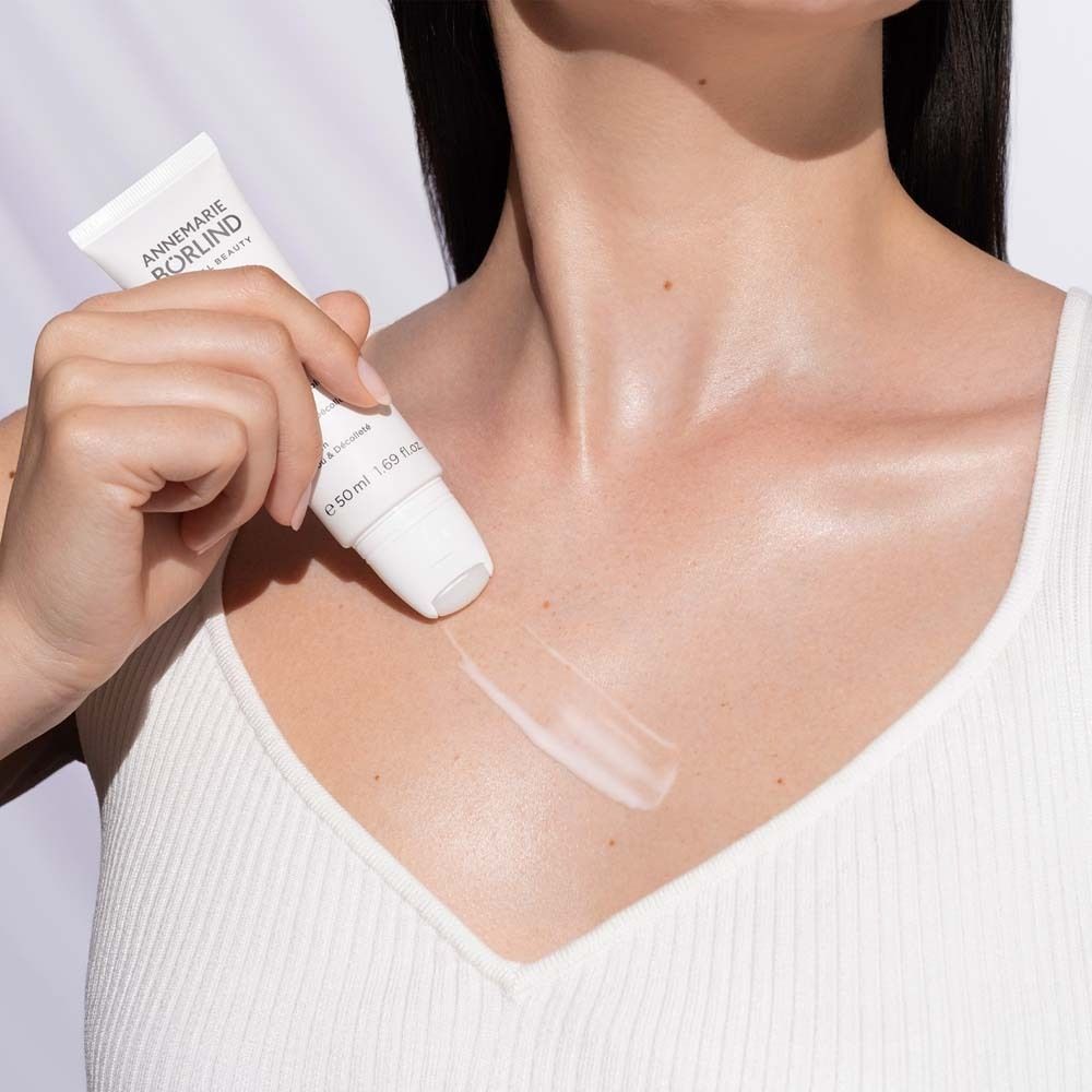 Frau trägt weißes Top. Sie hält eine Tube und trägt Serum auf den Hals auf. Marke: ANNEMARIE BÖRLIND.