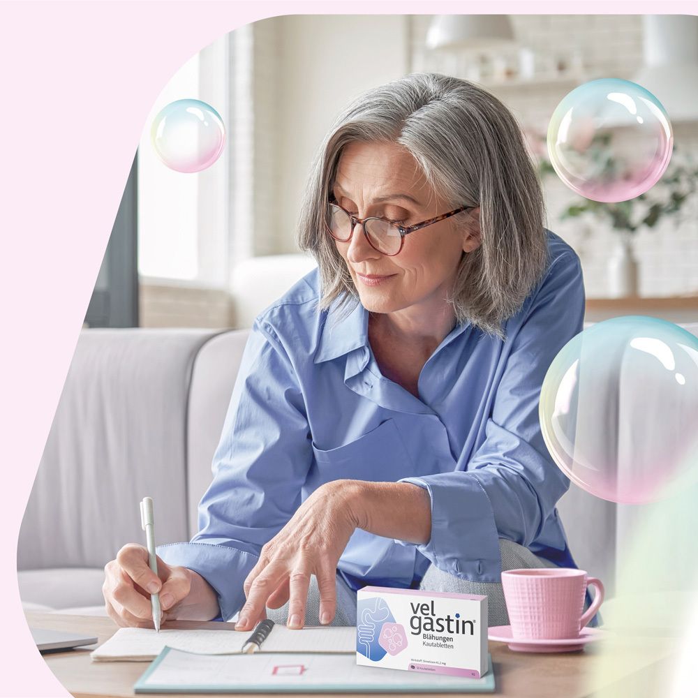 Frau am Schreibtisch mit Velgastin® Packung und Tasse. Zwei Tabletten liegen daneben.