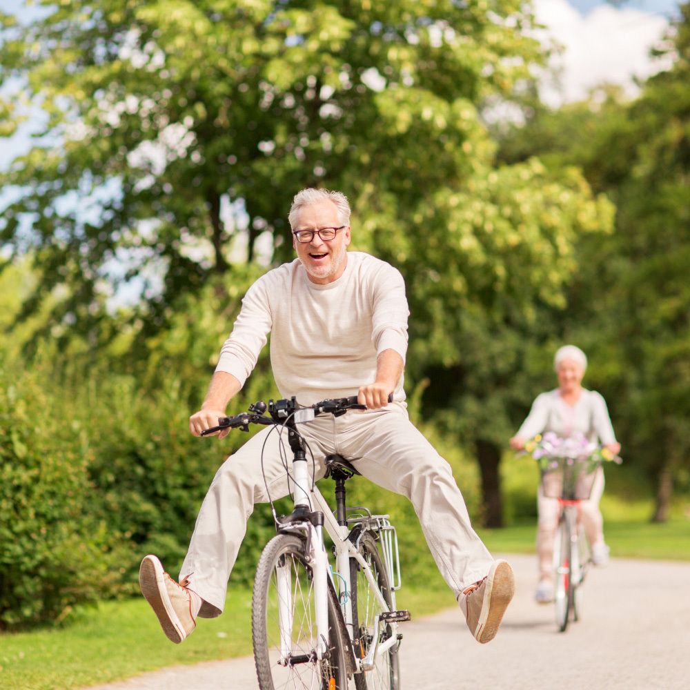 Zwei ältere Menschen fahren Fahrrad. Mann lacht. Frau fährt hinterher. Grüner Hintergrund.